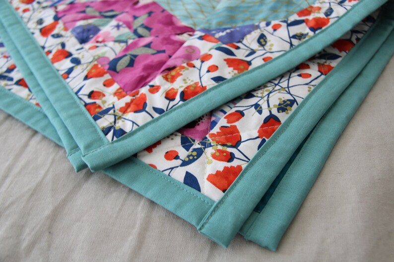 May include: Close-up of a quilted blanket with a floral pattern in pink, red, and blue on a white background. The blanket has a teal border. The quilt is folded over, showing the corner.