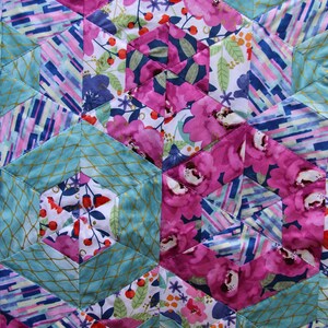 May include: Close-up of a quilted fabric with a geometric pattern. The design features hexagonal shapes in shades of pink, teal, and white. The pink sections display large floral patterns, while the teal sections have a gold net design.