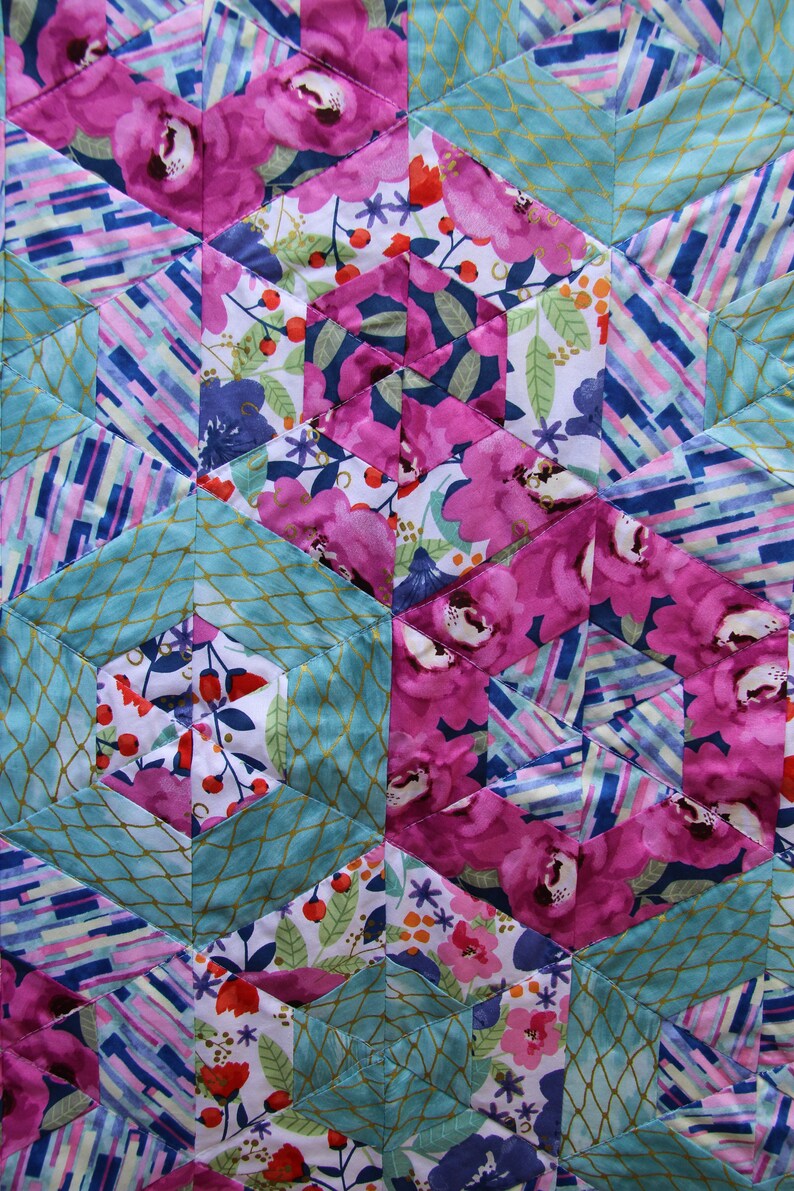 May include: Close-up of a quilted fabric with a geometric pattern. The design features hexagonal shapes in shades of pink, teal, and white. The pink sections display large floral patterns, while the teal sections have a gold net design.