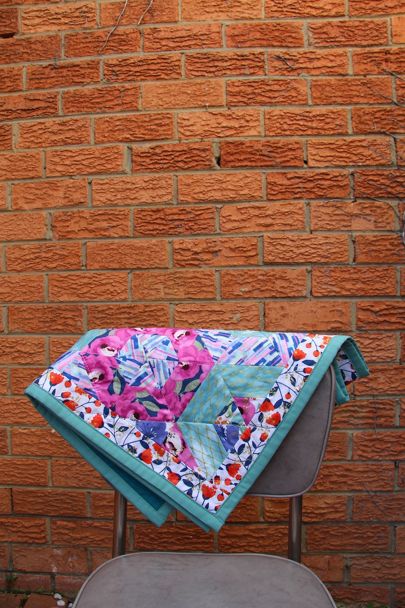 May include: A colorful quilt with a floral and geometric pattern, featuring pink flowers and blue accents. The quilt is bordered with a teal fabric and rests on a gray chair, set against a red brick wall.