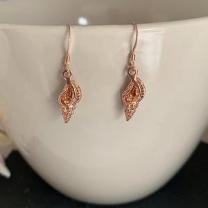 May include: A pair of rose gold seashell earrings. Each earring features a detailed, miniature conch shell design suspended from a simple hook. The earrings are displayed against a white background, highlighting their metallic sheen.