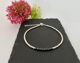 May include: A delicate bracelet featuring a continuous strand of small, polished silver-toned beads, with a short segment of dark, faceted charcoal-coloured beads integrated into the design, finished with a silver-toned lobster claw clasp.