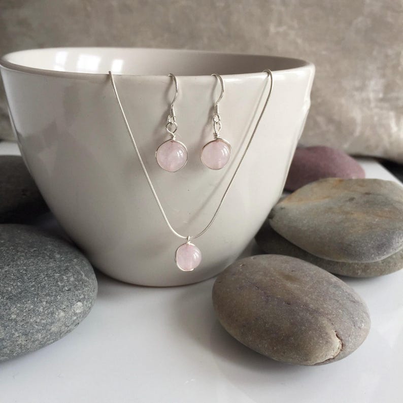 A silver necklace and earrings set with pink rose quartz stones. The necklace and earrings are displayed in a white bowl with grey stones.
