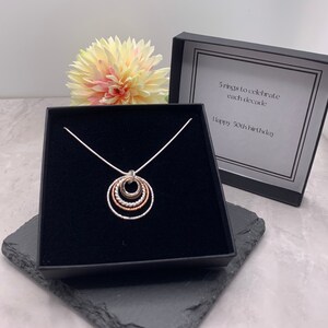 May include: A silver necklace with a pendant of five interlocking rings, each a different size and finish. The necklace is in a black box with a white label that reads "5 rings to celebrate each decade. Happy 50th birthday."