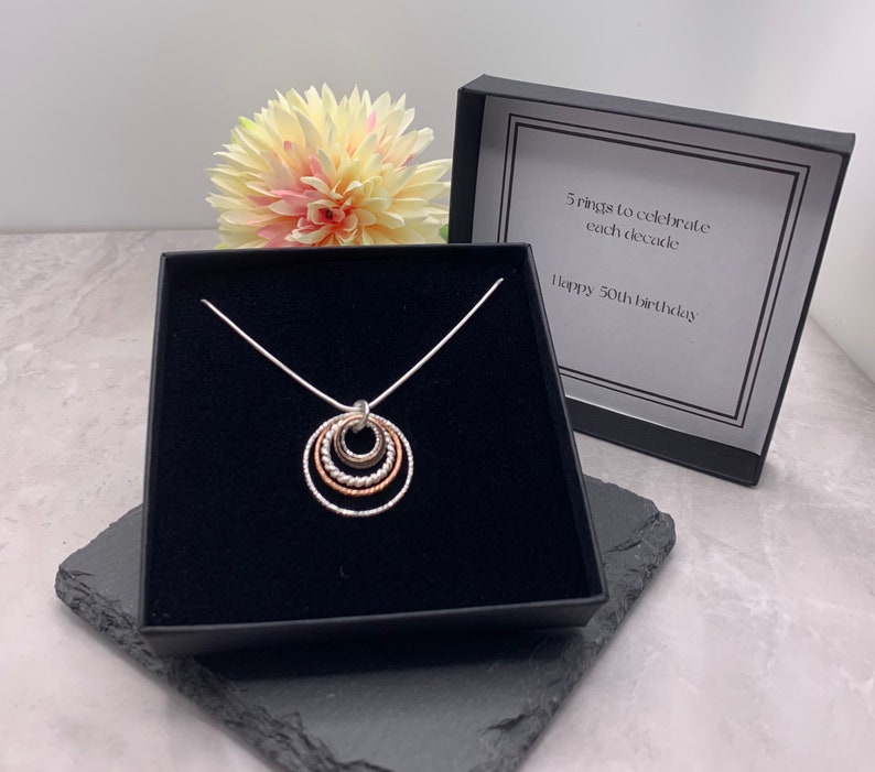 May include: A silver necklace with a pendant of five interlocking rings, each a different size and finish. The necklace is in a black box with a white label that reads "5 rings to celebrate each decade. Happy 50th birthday."