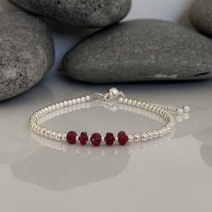 May include: A silver bracelet with a red gemstone accent. The bracelet is adjustable with a sliding clasp.