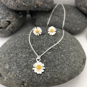 K&ouml;nnte beinhalten: Ein silbernes Halsketten- und Ohrring-Set mit einem zarten G&auml;nsebl&uuml;mchen-Design mit gelber Mitte. Die Halskette hat eine silberne Kette und einen kleinen G&auml;nsebl&uuml;mchen-Anh&auml;nger. Die Ohrringe sind klein und haben ein G&auml;nsebl&uuml;mchen-Design.