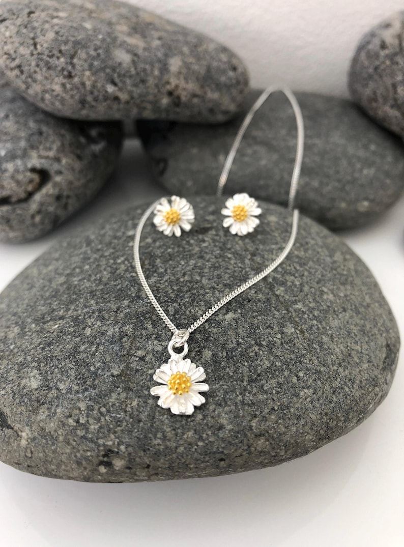K&ouml;nnte beinhalten: Ein silbernes Halsketten- und Ohrring-Set mit einem zarten G&auml;nsebl&uuml;mchen-Design mit gelber Mitte. Die Halskette hat eine silberne Kette und einen kleinen G&auml;nsebl&uuml;mchen-Anh&auml;nger. Die Ohrringe sind klein und haben ein G&auml;nsebl&uuml;mchen-Design.