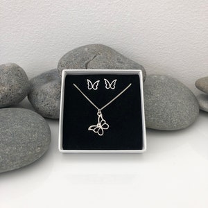 May include: A silver butterfly necklace and matching stud earrings set displayed in a white box with a black interior. The jewelry is set against a backdrop of gray stones and a white surface.
