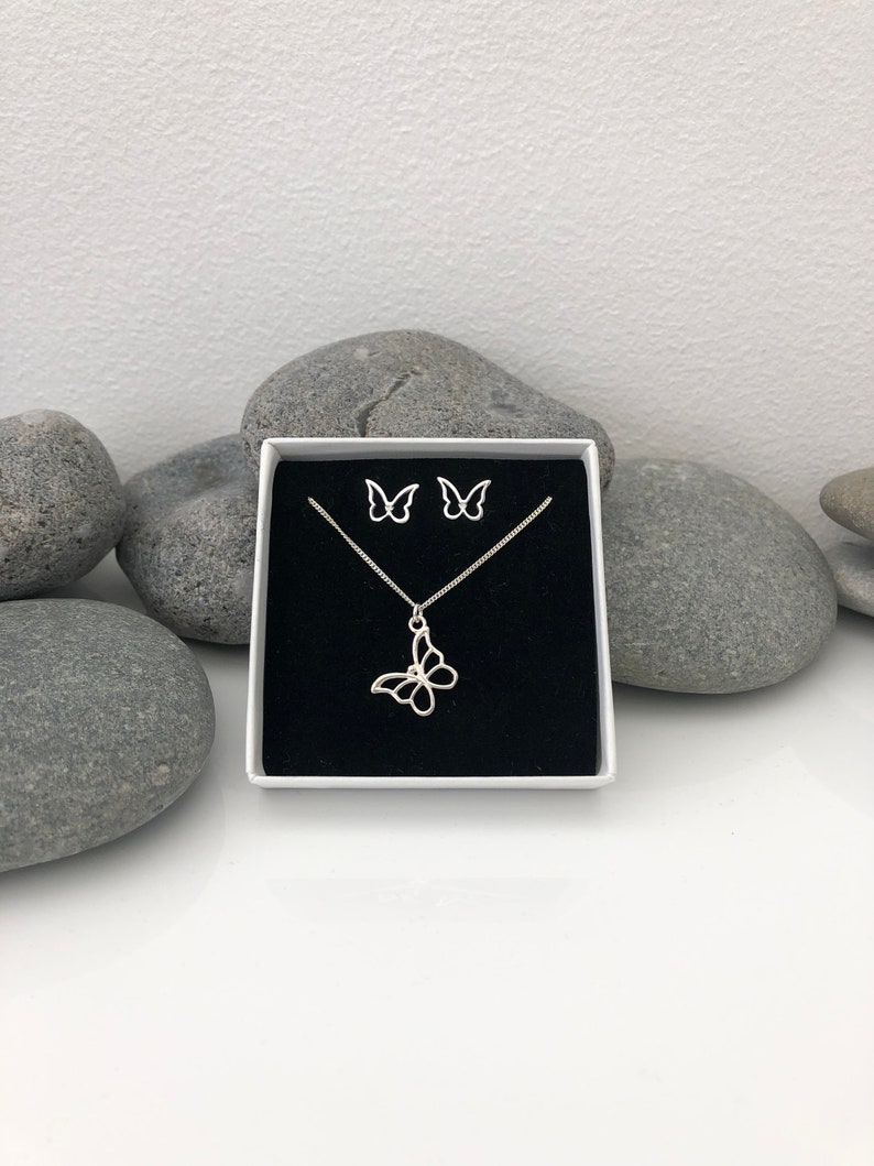 May include: A silver butterfly necklace and matching stud earrings set displayed in a white box with a black interior. The jewelry is set against a backdrop of gray stones and a white surface.