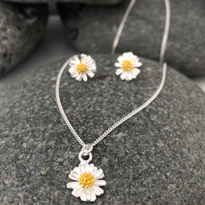K&ouml;nnte beinhalten: Ein silbernes Halsketten- und Ohrring-Set mit einem zarten G&auml;nsebl&uuml;mchen-Design. Die G&auml;nsebl&uuml;mchen haben eine gelbe Mitte und wei&szlig;e Bl&uuml;tenbl&auml;tter. Die Halskette ist eine einfache Kette mit einem Anh&auml;nger in Form eines G&auml;nsebl&uuml;mchens.