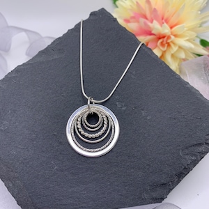 Pu&ograve; includere: Una collana d'argento con un ciondolo che presenta cinque cerchi concentrici, ognuno con un bordo testurizzato diverso. La collana &egrave; esposta su una superficie di ardesia nera.