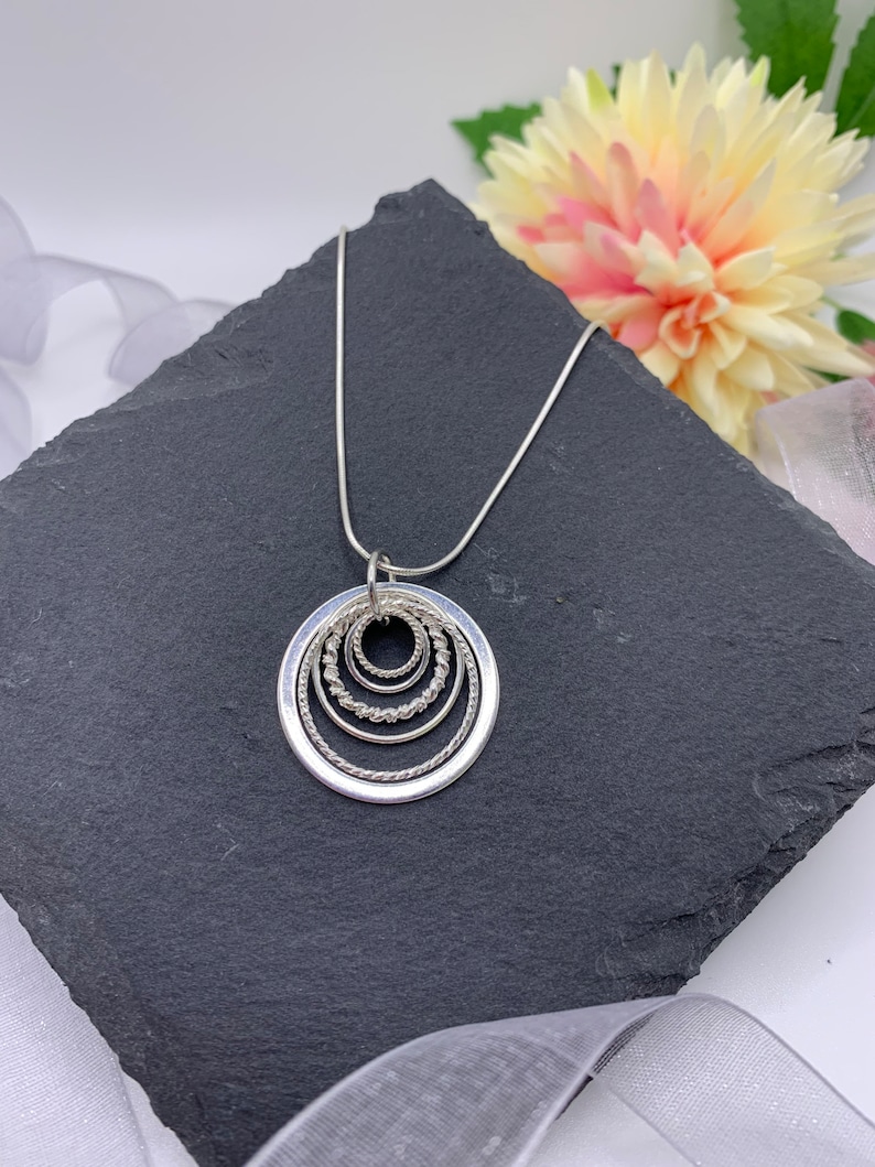 Pu&ograve; includere: Una collana d'argento con un ciondolo che presenta cinque cerchi concentrici, ognuno con un bordo testurizzato diverso. La collana &egrave; esposta su una superficie di ardesia nera.