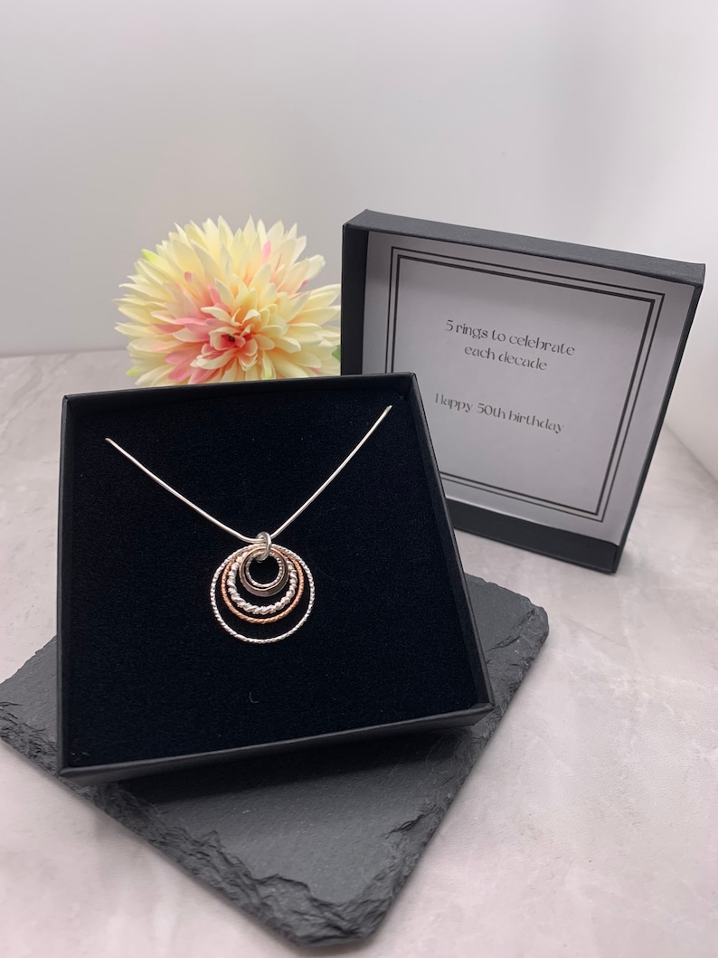 May include: A silver necklace with a pendant of five interlocking circles in silver, rose gold, and gold. The necklace is in a black box with a white card that reads "5 rings to celebrate each decade. Happy 50th birthday."
