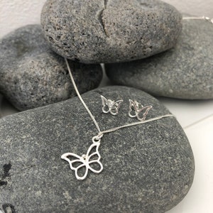 May include: A silver butterfly pendant necklace and matching stud earrings. The jewelry is displayed on a gray stone surface. The butterfly designs are open-work, showcasing the delicate shape. The necklace chain is thin and silver.