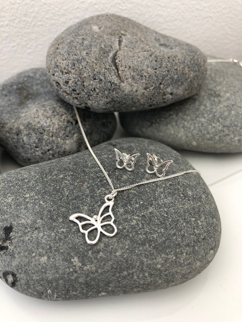 May include: A silver butterfly pendant necklace and matching stud earrings. The jewelry is displayed on a gray stone surface. The butterfly designs are open-work, showcasing the delicate shape. The necklace chain is thin and silver.