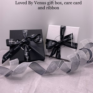 May include: Two gift boxes with black ribbon and the words "Loved By Venus" printed on them. The boxes are sitting on a white surface with a white and black ribbon.