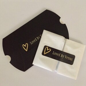 May include: A white tissue paper envelope with a black label that says "Loved By Venus" and a gold heart. The envelope is sitting on top of a black paper bag with a gold heart and the text "Loved By V" on it.