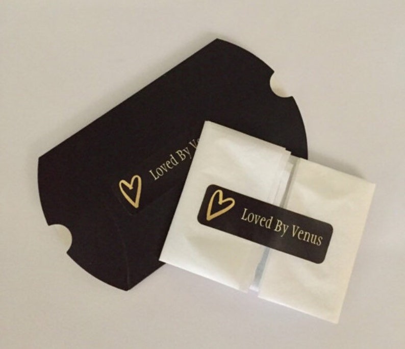 May include: A white tissue paper envelope with a black label that says "Loved By Venus" and a gold heart. The envelope is sitting on top of a black paper bag with a gold heart and the text "Loved By V" on it.
