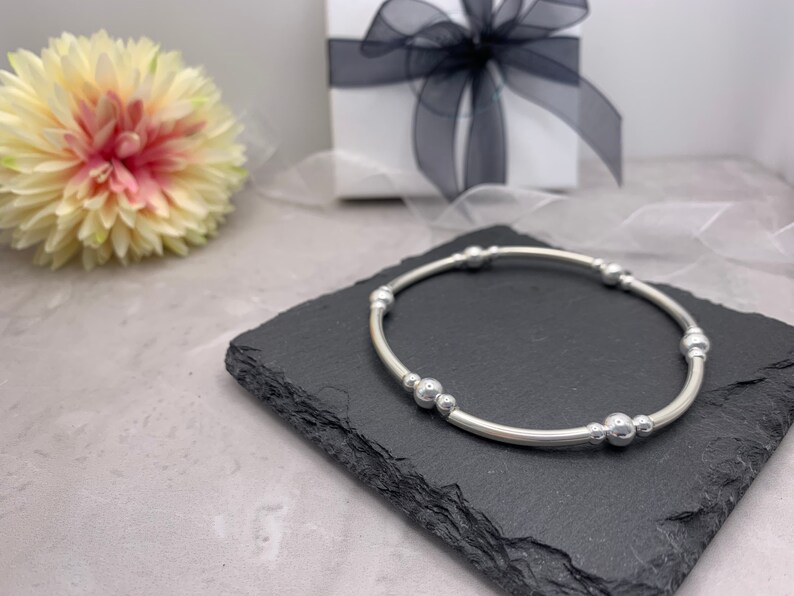 Puede incluir: Un brazalete de plata con una banda redonda lisa y cinco cuentas de plata. El brazalete est&aacute; sobre una superficie de pizarra negra.
