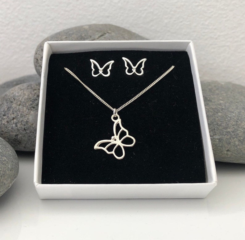 May include: A silver butterfly necklace and matching stud earrings set displayed in a white gift box with a black velvet interior. The butterfly pendant and earrings have an openwork design.