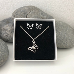 May include: A silver butterfly jewelry set, including a necklace and matching earrings, presented in a white box with a black interior. The butterfly pendant and earrings have an openwork design. The set is displayed against a backdrop of gray stones.
