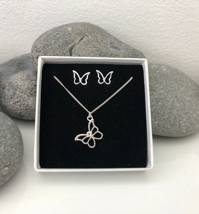 May include: A silver butterfly jewelry set, including a necklace and matching earrings, presented in a white box with a black interior. The butterfly pendant and earrings have an openwork design. The set is displayed against a backdrop of gray stones.