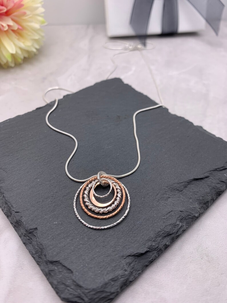 May include: A silver chain necklace with a pendant made of four interlocking circles. The circles are made of silver and rose gold, and they are arranged in a graduated size.