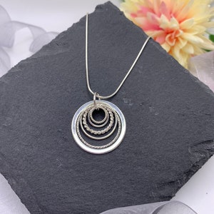 Pu&ograve; includere: Una collana d'argento con un ciondolo che presenta cinque cerchi concentrici, ognuno con un bordo testurizzato diverso. La collana &egrave; esposta su una superficie di ardesia nera.
