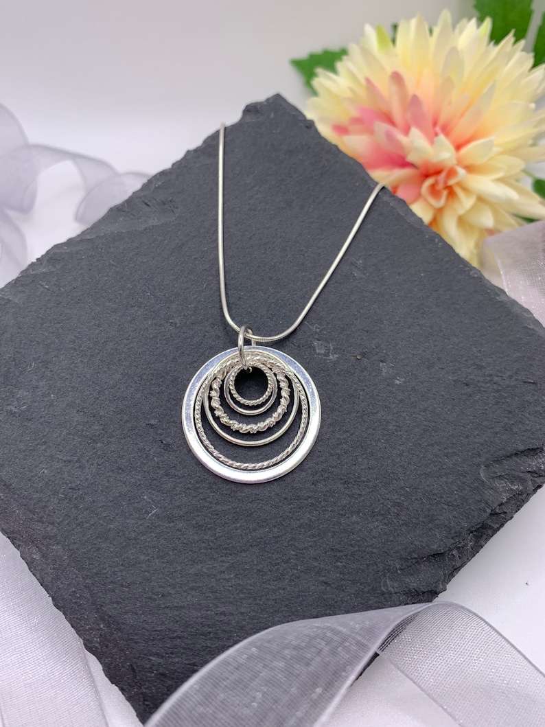 Pu&ograve; includere: Una collana d'argento con un ciondolo che presenta cinque cerchi concentrici, ognuno con un bordo testurizzato diverso. La collana &egrave; esposta su una superficie di ardesia nera.