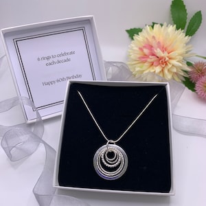 Pu&ograve; includere: Una collana d'argento con un ciondolo composto da sei cerchi intrecciati. La collana &egrave; in una scatola bianca con interno nero. La scatola contiene un biglietto che recita "6 anelli per celebrare ogni decennio Buon 60&deg; compleanno".