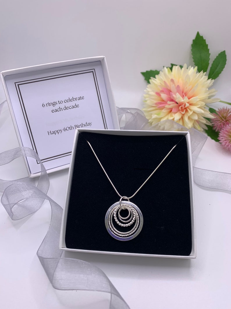 Pu&ograve; includere: Una collana d'argento con un ciondolo composto da sei cerchi intrecciati. La collana &egrave; in una scatola bianca con interno nero. La scatola contiene un biglietto che recita "6 anelli per celebrare ogni decennio Buon 60&deg; compleanno".