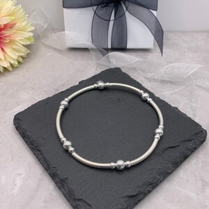 Puede incluir: Un brazalete plateado con peque&ntilde;as cuentas redondas. El brazalete est&aacute; sobre una superficie de pizarra negra.