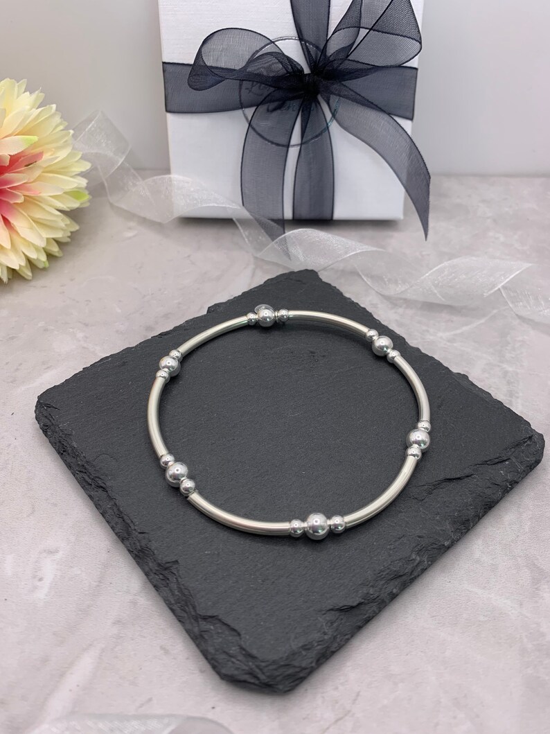 Puede incluir: Un brazalete plateado con peque&ntilde;as cuentas redondas. El brazalete est&aacute; sobre una superficie de pizarra negra.