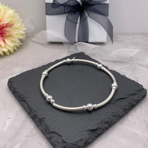 Puede incluir: Un brazalete plateado con una banda fina y peque&ntilde;as cuentas redondas. El brazalete est&aacute; sobre una superficie de pizarra negra.