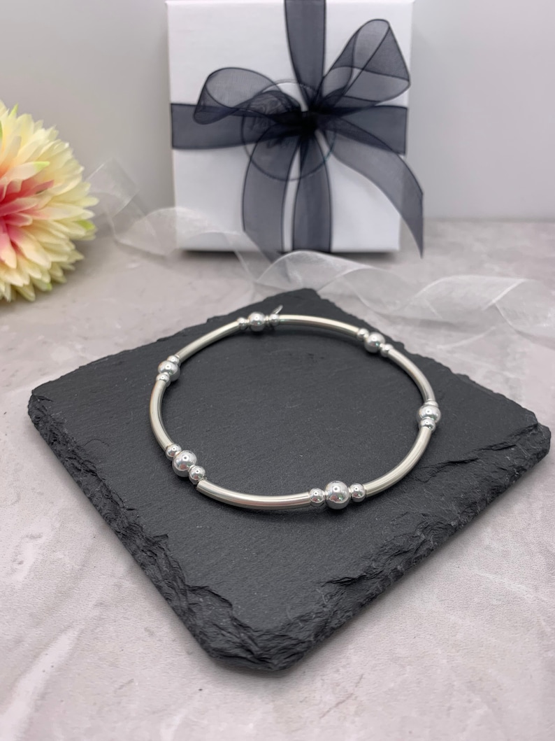 Puede incluir: Un brazalete plateado con una banda fina y peque&ntilde;as cuentas redondas. El brazalete est&aacute; sobre una superficie de pizarra negra.