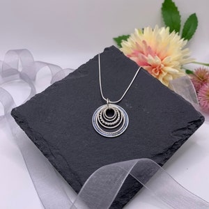 Pu&ograve; includere: Una collana d'argento con un ciondolo che presenta cinque cerchi concentrici, ognuno con un bordo testurizzato diverso. I cerchi sono d'argento con una riempitura in smalto blu.