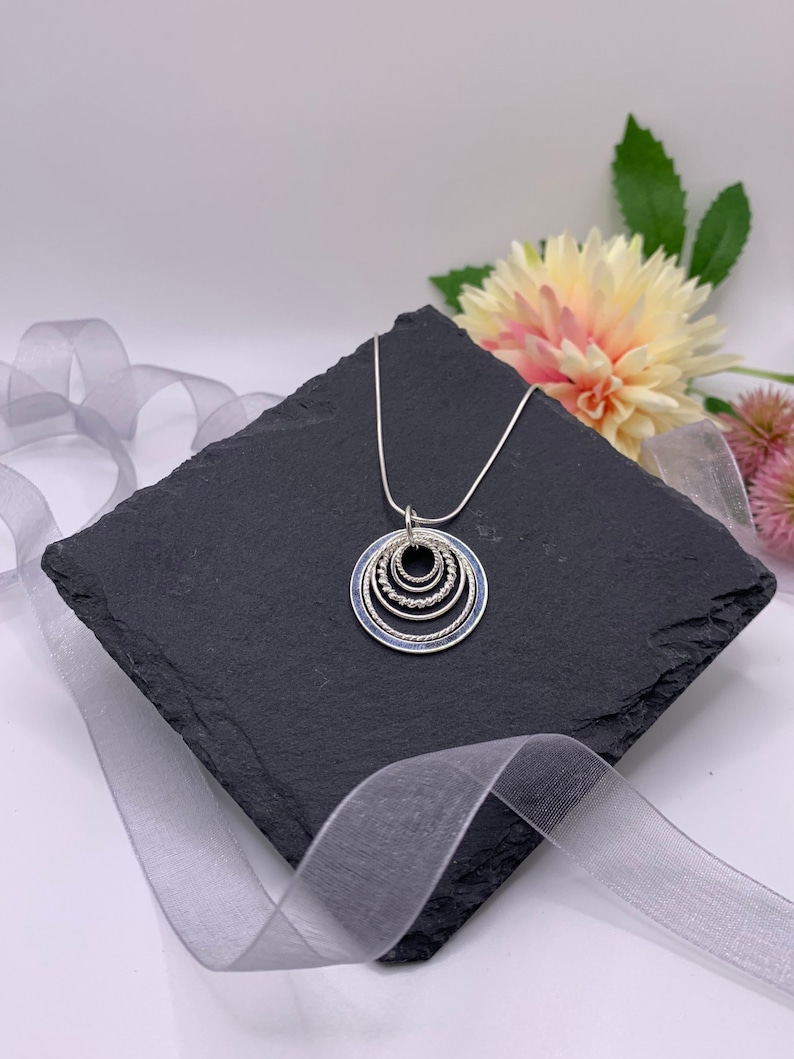 Pu&ograve; includere: Una collana d'argento con un ciondolo che presenta cinque cerchi concentrici, ognuno con un bordo testurizzato diverso. I cerchi sono d'argento con una riempitura in smalto blu.