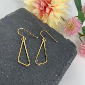 May include: Two gold-coloured, open-frame triangle earrings with hook closures. The earrings are presented on a dark grey, textured surface, with soft-focus flowers in the background.
