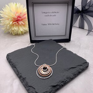 May include: A silver necklace with a pendant of five interlocking circles. The circles are made of silver and rose gold. The necklace is displayed on a black slate tile. The box in the background has a card that reads "5 rings to celebrate each decade. Happy 50th birthday."