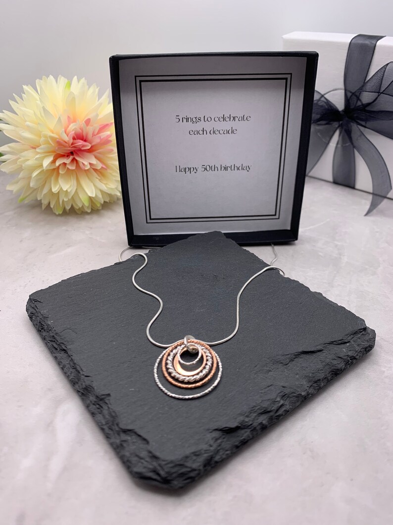 May include: A silver necklace with a pendant of five interlocking circles. The circles are made of silver and rose gold. The necklace is displayed on a black slate tile. The box in the background has a card that reads "5 rings to celebrate each decade. Happy 50th birthday."