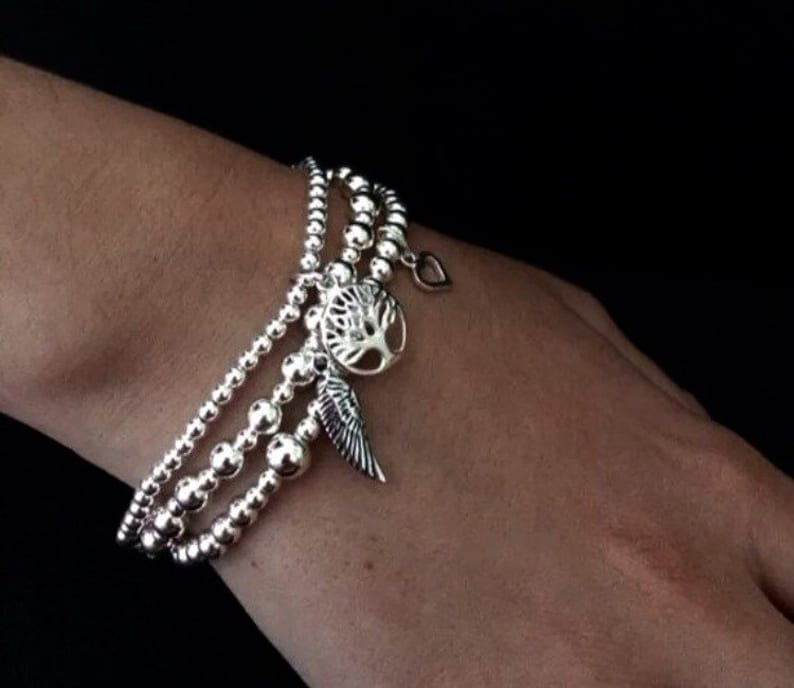 May include: A silver bracelet with three strands of beads and three charms: a tree, a heart, and a wing.