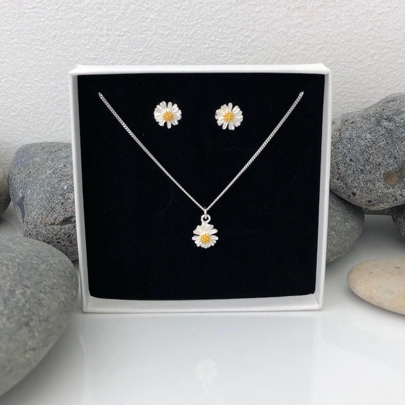 K&ouml;nnte beinhalten: Ein silbernes Halsketten- und Ohrring-Set mit einem zarten G&auml;nsebl&uuml;mchen-Design mit gelber Mitte. Der Schmuck ist in einer schwarzen Samtbox mit wei&szlig;em Deckel ausgestellt.