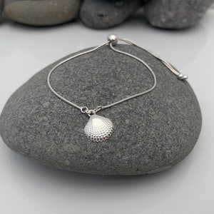 Könnte beinhalten: Ein verstellbares silbernes Armband mit einem kleinen silbernen Muschel-Anhänger.