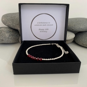 May include: A silver bracelet with six red gemstones, displayed in a black gift box. The box's interior has text saying, "6 birthstones to celebrate each decade" and "Happy 60th birthday."