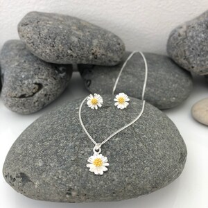 K&ouml;nnte beinhalten: Ein silbernes Halsketten- und Ohrring-Set mit einem zarten G&auml;nsebl&uuml;mchen-Design mit goldener Mitte. Der Schmuck wird auf einem grauen Steinuntergrund pr&auml;sentiert.