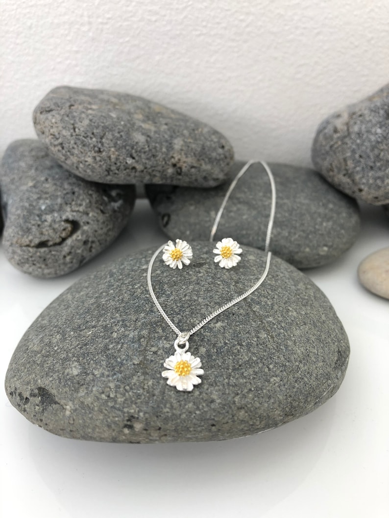 K&ouml;nnte beinhalten: Ein silbernes Halsketten- und Ohrring-Set mit einem zarten G&auml;nsebl&uuml;mchen-Design mit goldener Mitte. Der Schmuck wird auf einem grauen Steinuntergrund pr&auml;sentiert.