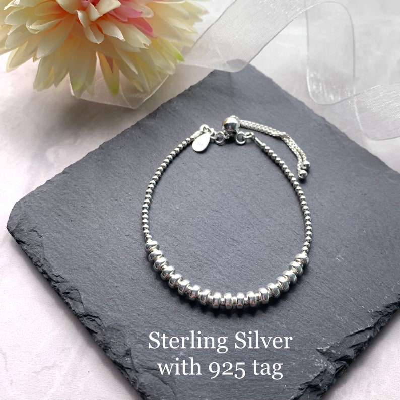 A silver beaded bracelet with a sliding clasp and a 925 tag. The bracelet is on a grey stone surface.