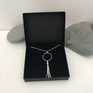 May include: A silver necklace with a circle pendant and a chain tassel. The necklace is in a black box.
