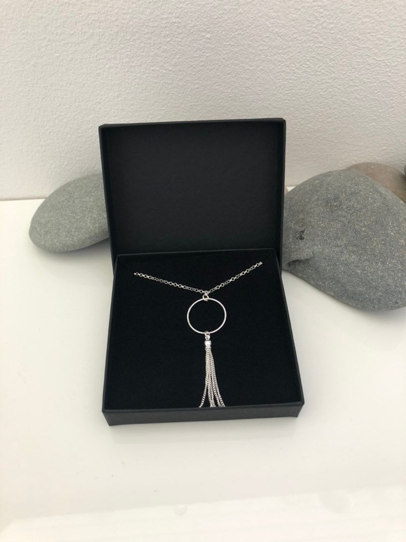 May include: A silver necklace with a circle pendant and a chain tassel. The necklace is in a black box.